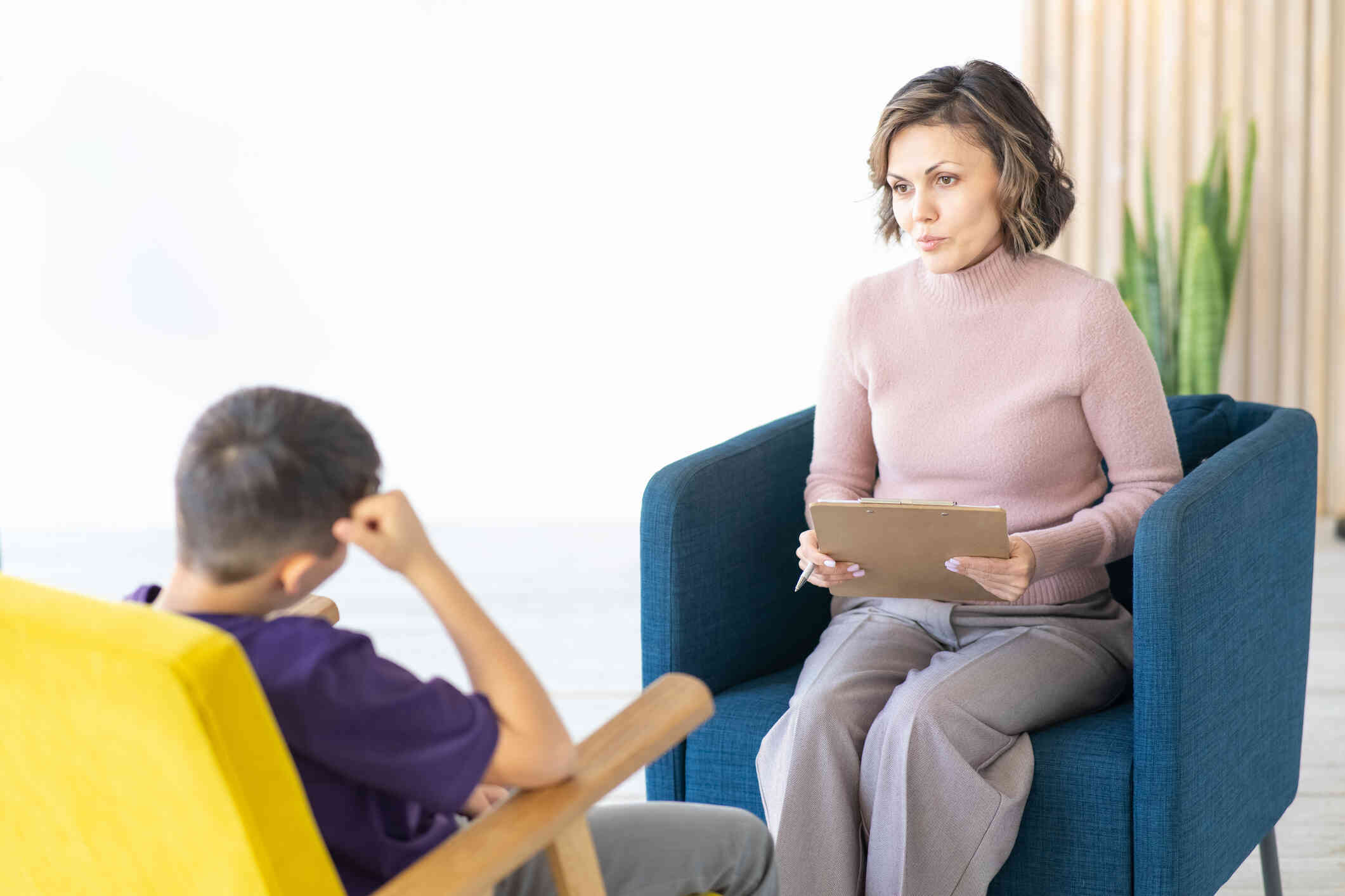 A young boy sits in a yellow shair and listens to the female therapist sitting across from him during a therapy session.
